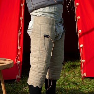 May include: A pair of beige quilted leg armor with leather straps and buckles. The armor is worn over a white undergarment and is visible against a red background.