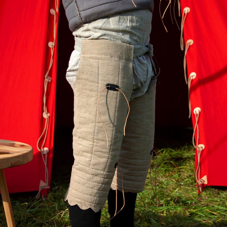 May include: A pair of beige quilted leg armor with leather straps and buckles. The armor is worn over a white undergarment and is visible against a red background.