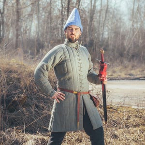 May include: A person wearing a light blue conical hat and a quilted, olive-green tunic with a brown belt and a red sword. The outfit is reminiscent of medieval attire. The sword has a red leather handle.