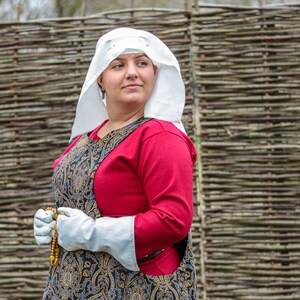 May include: A woman wearing a white linen head covering and a red long-sleeved tunic with a black and gold patterned apron. She is holding a string of amber beads in her hands.