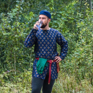May include: A man wearing a blue patterned shirt with gold accents, a blue hat, and a red belt with a green glove attached. He is standing in a woodland setting.