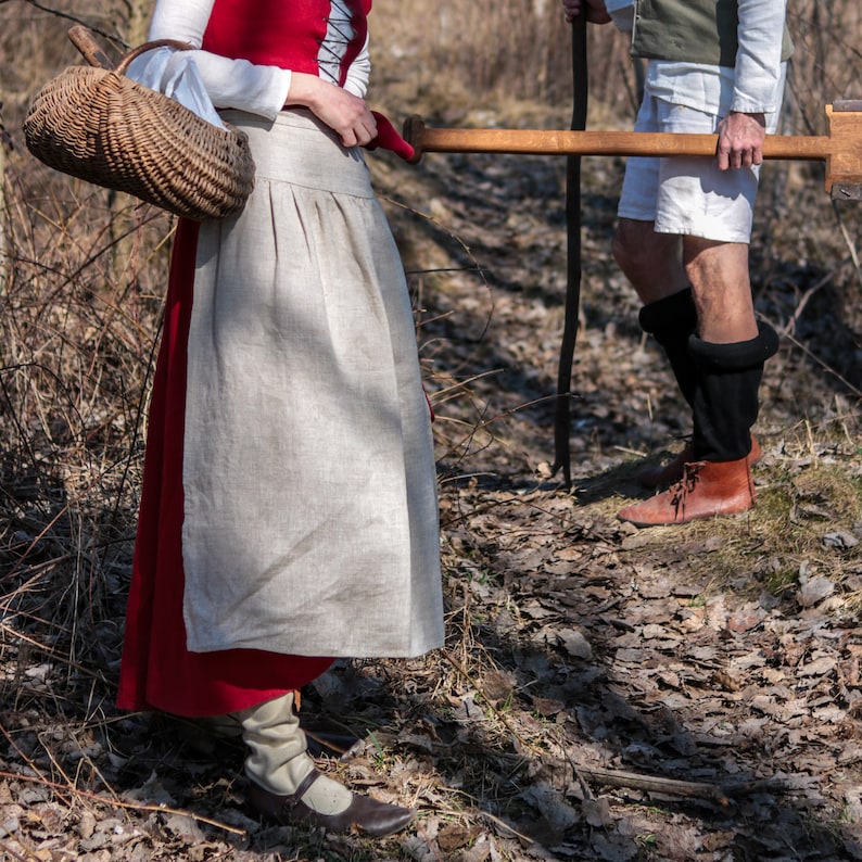 Puede incluir: Una mujer con un vestido rojo y blanco lleva una cesta de mimbre. Camina por un bosque con un hombre que lleva un bast&oacute;n de madera.