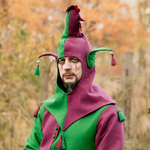 Op de afbeelding: Een tweekleurige kap van groene en paarse viltstof in de vorm van een haan met een rode kam en lellen. De kap is voorzien van twee zijpunten met kleine zilveren belletjes en bijpassende kwastjes, en heeft een knoopsluiting aan de voorzijde.
