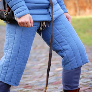 Op de afbeelding: Blauwe gewatteerde broek met een bruine leren riem en een lange band die naar beneden hangt. De broek wordt gedragen door een persoon en heeft een gewatteerd ontwerp.
