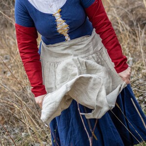 Puede incluir: Una mujer con un vestido medieval azul y rojo con un delantal de lino. El delantal ondea con el viento y la mujer lo sujeta con su mano derecha.