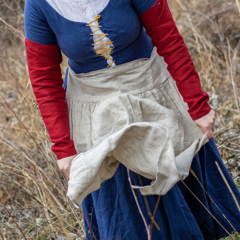 Puede incluir: Una mujer con un vestido medieval azul y rojo con un delantal de lino. El delantal ondea con el viento y la mujer lo sujeta con su mano derecha.