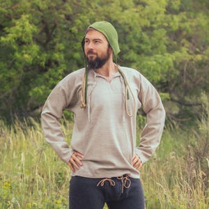 Op de afbeelding: Een beige tuniek met lange mouwen en een V-hals, gecombineerd met een donkergrijze broek met een trekkoord in de taille. Een groene kap met lange, dikke beige touwbanden hangt om de nek en rust over de schouders van de tuniek.