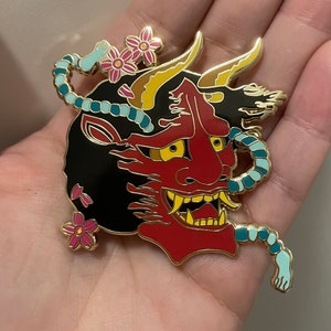 May include: A red, black, and gold enamel pin featuring a Japanese Oni mask design. The pin has yellow eyes, a fierce expression, and is adorned with pink cherry blossoms and a blue rope-like detail.