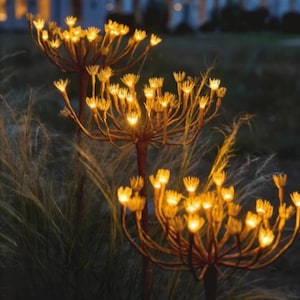 May include: Outdoor decorative metal flower sculptures with warm yellow lights. The sculptures have a brown stem and multiple branches with small, glowing flower-like lights at the ends. The background features tall grass and a blurred night scene.