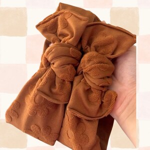 May include: Two rust-colored headbands with a knotted bow design. The fabric has a textured floral pattern. The headbands are held in a hand against a checkered background of beige, pink, and white squares.
