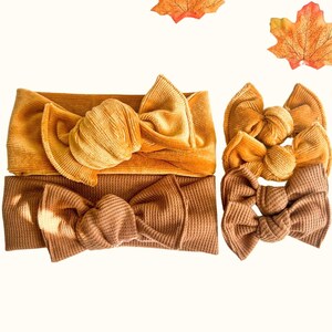 May include: A selection of headbands in gold and brown hues, each with a large bow. The arrangement includes three decorative orange and yellow autumn leaves. The headbands are made from corduroy and waffle knit fabrics.