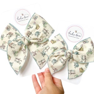May include: Two handmade hair bows with a light green and white fabric featuring a whimsical print of bugs and butterflies. The bows are tied with a light blue ribbon and have a small white circle label with the text "Koko-Bee - Handcrafted Bows".