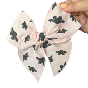 May include: A large, light pink hair bow with a repeating pattern of gray bats and white stars. The bow is held by a hand with a light orange nail polish. The edges of the bow are finished with a black zigzag stitch.