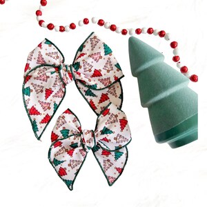 May include: Two Christmas tree patterned hair bows with green trim. The larger bow is tied in a traditional bow shape, while the smaller bow is tied in a more modern style.