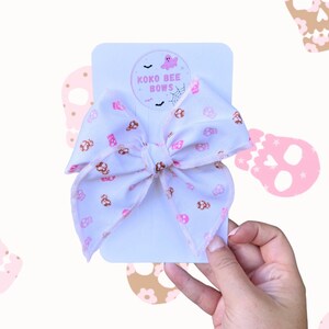 May include: A white bow with a pink and brown skull print. The bow is tied with a white ribbon and has a small white tag with the text "Koko Bee Bows" in pink letters.