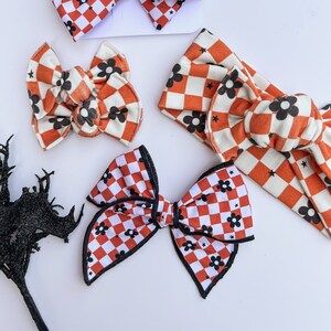 May include: A collection of hair accessories featuring a red and white checkered pattern with black flower and star accents. The set includes a large bow, a smaller bow, a headband, and a bow clip. The accessories are on a white background.
