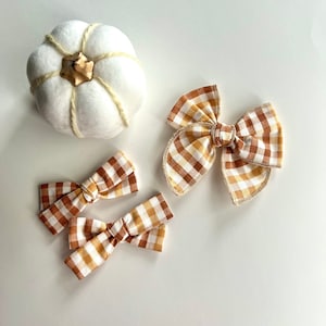 May include: An overhead shot features a white pumpkin and three hair bows. The bows have a brown, white, and yellow plaid pattern. The pumpkin is white with a brown stem and beige twine accents. The bows are in various sizes.