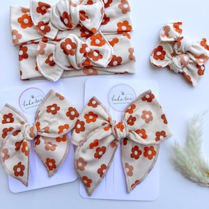 May include: A collection of baby headbands and hair bows. The headbands and bows are cream-colored with a repeating pattern of orange flowers. The bows are attached to white cards with the text "koko bee handcrafted boutique".