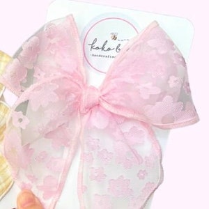 May include: A large, pink, floral-patterned hair bow made of sheer fabric. The bow is attached to a white card with a circular logo that reads "koko b handcrafted". The bow is held in place by a hand.