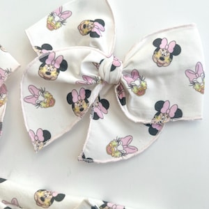 May include: A white fabric bow with a pink and white Minnie Mouse print. The bow is tied in a knot and has a pink and white polka dot pattern.