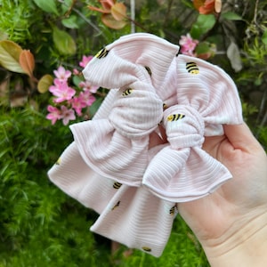 May include: Two pink hair bows with a bee pattern. The bows are made of a soft, ribbed fabric.