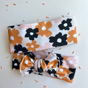 Op de afbeelding: Twee haarbanden met een bloemenpatroon. Het patroon heeft grote bloemen in zwart, bruin en roze op een witte achtergrond. E&eacute;n haarband is een eenvoudige band, de andere heeft een strik. Kleine oranje sprinkles zijn verspreid.