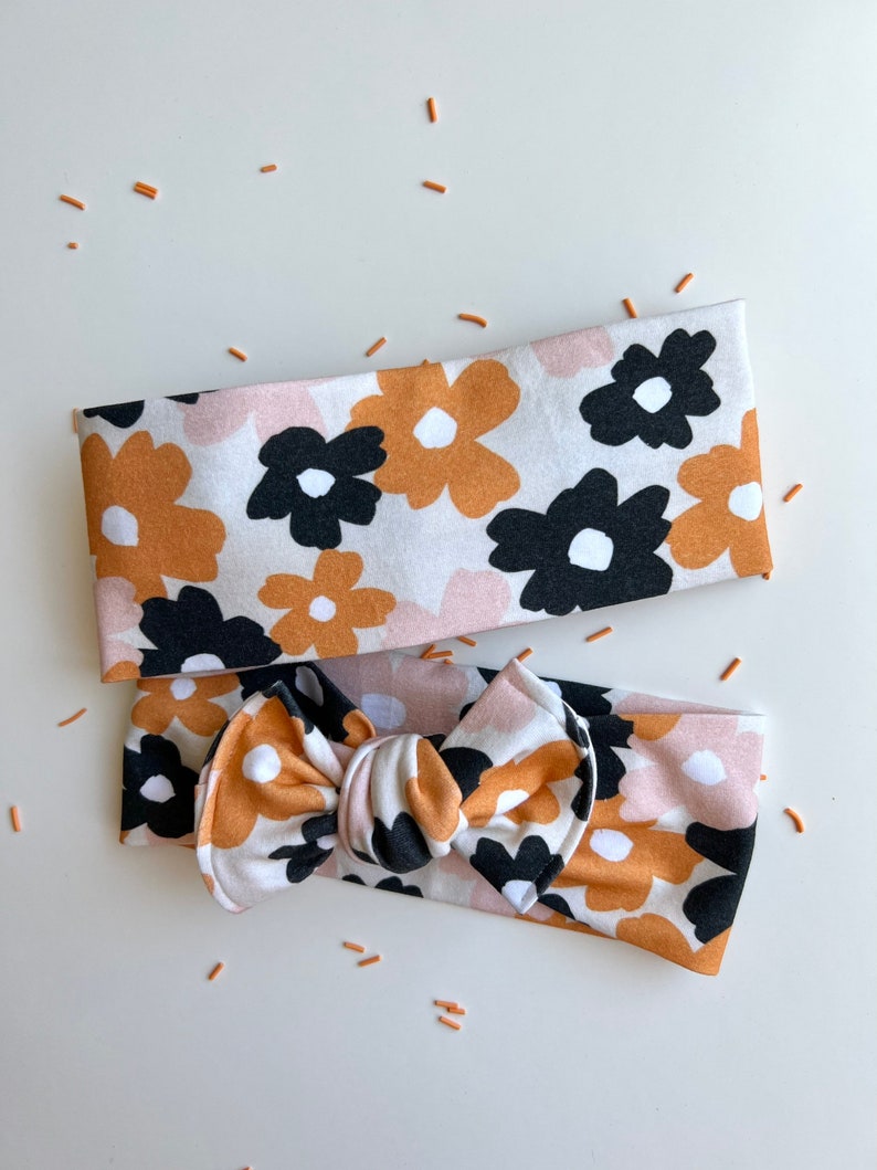 Op de afbeelding: Twee haarbanden met een bloemenpatroon. Het patroon heeft grote bloemen in zwart, bruin en roze op een witte achtergrond. E&eacute;n haarband is een eenvoudige band, de andere heeft een strik. Kleine oranje sprinkles zijn verspreid.