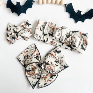 May include: A set of Halloween-themed hair accessories, including a large bow, a smaller bow, and a headband. The accessories feature a cream-colored fabric with a pattern of black bats and orange floral accents. A bat garland hangs above.