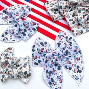 May include: Four red, white, and blue Mickey Mouse and Minnie Mouse patterned hair bows. The bows are made of fabric and have a white background with red, white, and blue accents. The bows are arranged on a white surface.