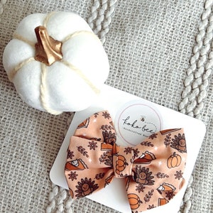 May include: A peach-colored bow with a Thanksgiving-themed print featuring turkeys, pumpkins, and pie slices. The bow is on a white card with a logo that says "Koko Bee - Handcrafted Bows".