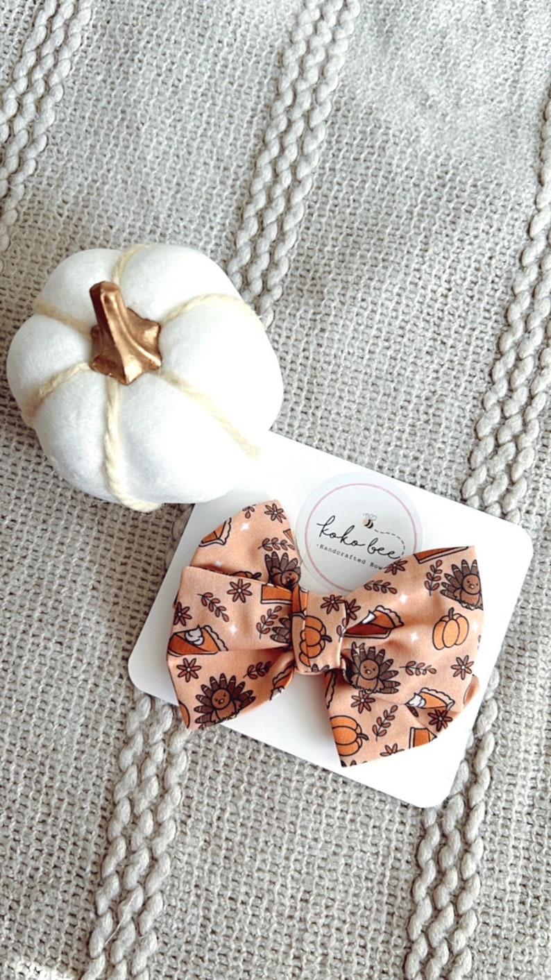 May include: A peach-colored bow with a Thanksgiving-themed print featuring turkeys, pumpkins, and pie slices. The bow is on a white card with a logo that says "Koko Bee - Handcrafted Bows".