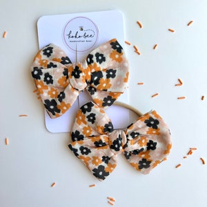Op de afbeelding: Twee bloemenstrikken en een bijpassende haarband. De strikken hebben een bruinachtige stof met een patroon van zwarte en oranje bloemen. De strikken zijn bevestigd aan een witte kaart met de tekst "koko-bee - handcrafted bows-".