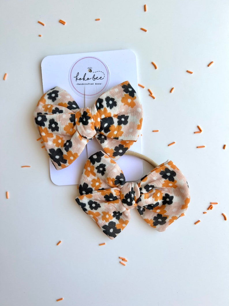 Op de afbeelding: Twee bloemenstrikken en een bijpassende haarband. De strikken hebben een bruinachtige stof met een patroon van zwarte en oranje bloemen. De strikken zijn bevestigd aan een witte kaart met de tekst "koko-bee - handcrafted bows-".