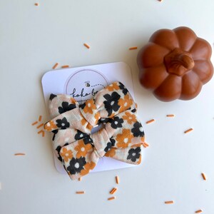 Op de afbeelding: Een haarstrik met bloemenpatroon in perzik, zwart en goud, met een pompoendecoratie. De strik zit op een witte kaart met het logo "koko bee handcrafted". Oranje sprinkles zijn rond de strik verspreid.