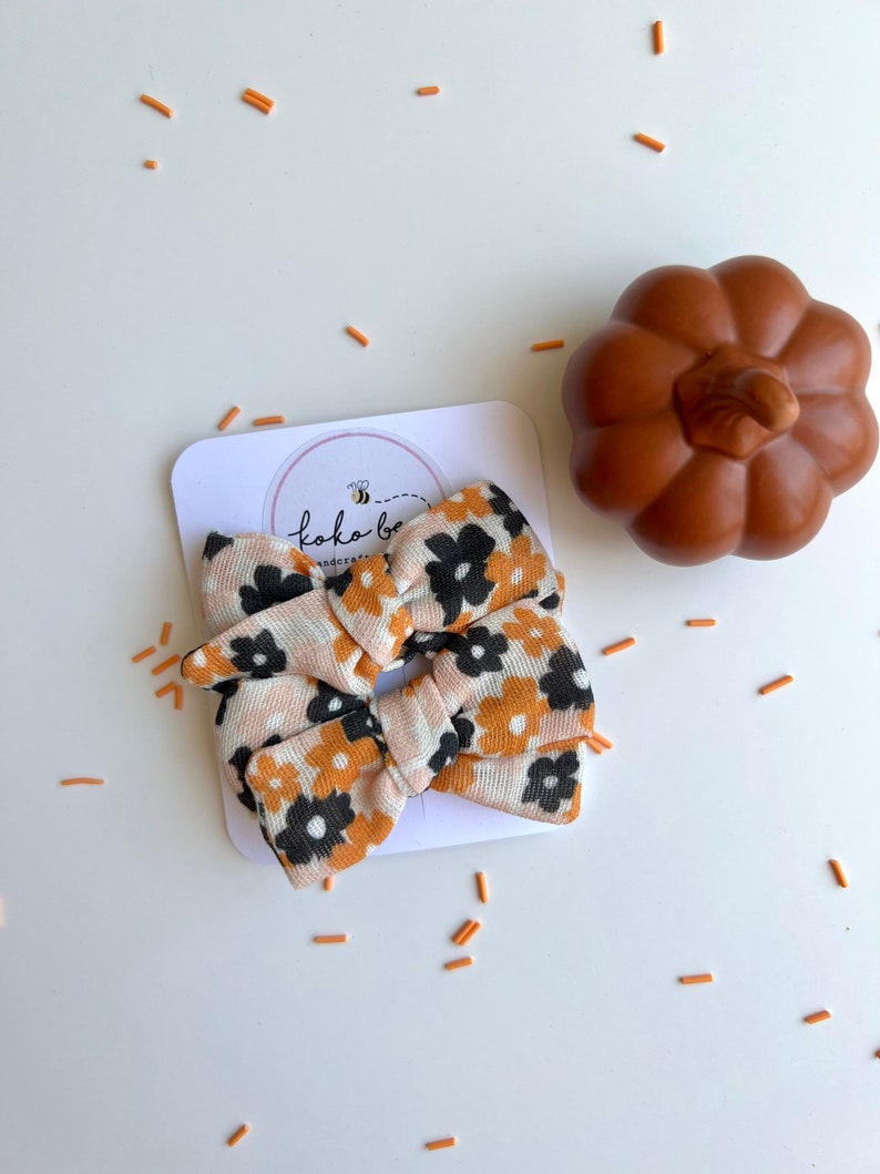 Op de afbeelding: Een haarstrik met bloemenpatroon in perzik, zwart en goud, met een pompoendecoratie. De strik zit op een witte kaart met het logo "koko bee handcrafted". Oranje sprinkles zijn rond de strik verspreid.