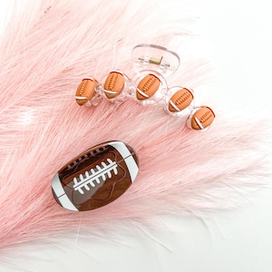 May include: A clear plastic hair claw clip adorned with miniature football-shaped embellishments. A brown and white football-shaped case is also visible. The items are set against a backdrop of pink feathers.