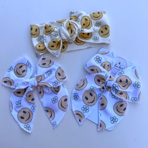 Pode incluir: Uma faixa de cabelo creme e dois la&ccedil;os de cabelo a condizer, todos adornados com um padr&atilde;o repetido de rostos sorridentes e pequenas flores de margarida em amarelo e castanho. Os la&ccedil;os t&ecirc;m um centro atado e caudas longas e esvoa&ccedil;antes.