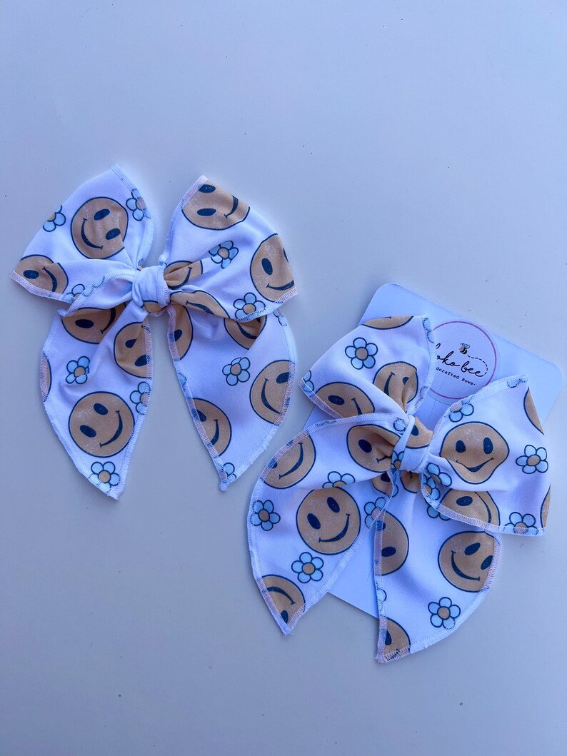 Pode incluir: Duas fitas de cabelo de tecido branco com um padr&atilde;o repetido de rostos sorridentes e pequenas flores de margarida em bege e preto. Uma fita &eacute; maior que a outra. A fita menor &eacute; presa a um cart&atilde;o branco com um logotipo.