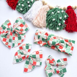 May include: A collection of hair bows and a headband with a red, green, and white checkered floral pattern. A Christmas-themed garland with red, green, white, and pink tassels and wooden beads is in the background.