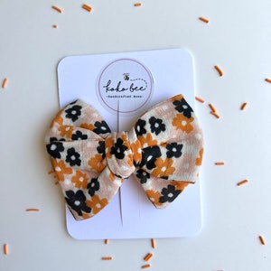 Op de afbeelding: Een haarstrik met bloemenpatroon in tinten beige, zwart en oranje, gepresenteerd op een witte kaart. De strik heeft een herhalend bloemenontwerp en is gelabeld met "koko bee - Handcrafted Bows."