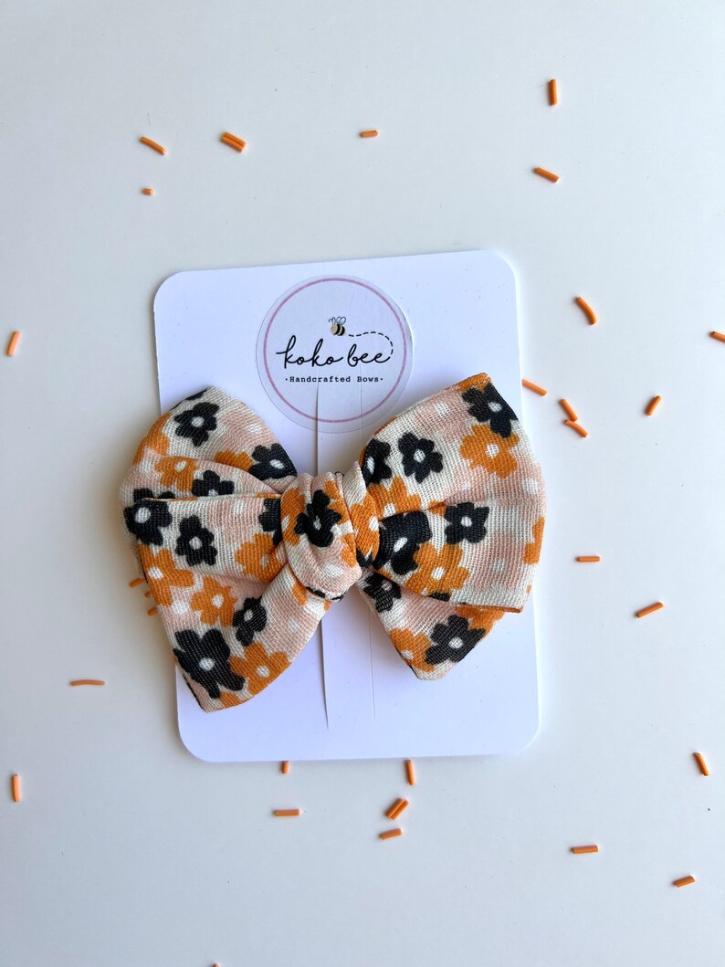 Op de afbeelding: Een haarstrik met bloemenpatroon in tinten beige, zwart en oranje, gepresenteerd op een witte kaart. De strik heeft een herhalend bloemenontwerp en is gelabeld met "koko bee - Handcrafted Bows."