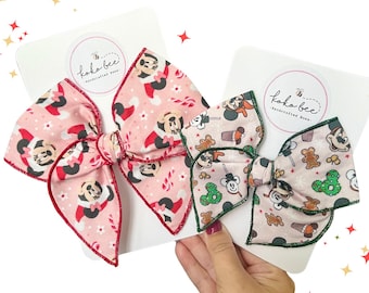 Puede incluir: Dos lazos para el pelo hechos a mano con diseños de Mickey Mouse y navideños. Uno es rosa con Mickey y bastones de caramelo, el otro es rosa claro con Mickey, hombres de jengibre y árboles de Navidad. Ambos están sobre tarjetas blancas.