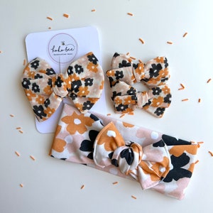 Op de afbeelding: Een set bloemen haaraccessoires, waaronder twee kleine strikken, een grote strik op een kaart en een haarband. De strikken en haarband hebben een patroon van zwarte, bruine en witte bloemen op een lichtroze achtergrond. De kaart heeft de tekst "koka bee - handcrafted bows."