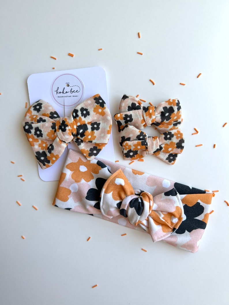 Op de afbeelding: Een set bloemen haaraccessoires, waaronder twee kleine strikken, een grote strik op een kaart en een haarband. De strikken en haarband hebben een patroon van zwarte, bruine en witte bloemen op een lichtroze achtergrond. De kaart heeft de tekst "koka bee - handcrafted bows."