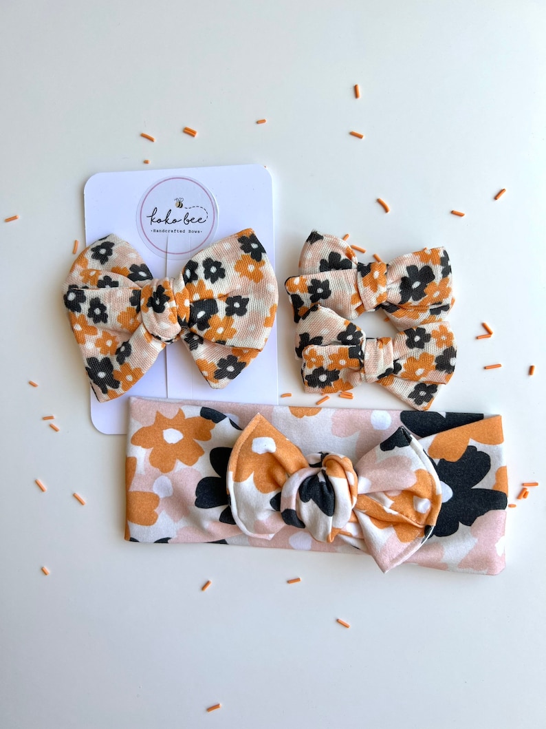 Op de afbeelding: Een set bloemen haaraccessoires, waaronder twee strikclips, een hoofdband en een grotere strikclip. De stof heeft een patroon van zwarte, bruine en witte bloemen. De accessoires worden gepresenteerd op een wit oppervlak met de merknaam "koko bee".