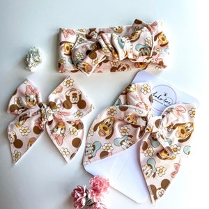 May include: A set of two pink and brown hair bows with a floral and cartoon character print. The bows are made of fabric and have a knotted center. The print features cartoon characters from Disney's Mickey Mouse and Minnie Mouse. The bows are displayed on a white background with a small white card that says "Lola Bee - handcrafted with love".