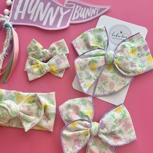 May include: A set of four floral hair bows with a purple trim. The bows are made of a white fabric with a floral print in shades of pink, yellow, and purple. The bows are arranged on a pink background with a purple banner that says "Hunny Bunny".