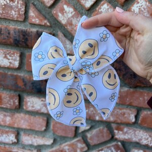 Pode incluir: Um la&ccedil;o de tecido azul claro com um padr&atilde;o repetido de rostos sorridentes amarelos e pequenas flores brancas. O la&ccedil;o &eacute; amarrado no centro e tem um formato cl&aacute;ssico, ideal para acess&oacute;rios de cabelo.