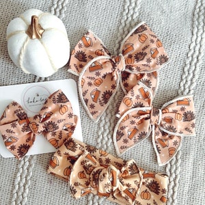 May include: Three brown and orange fabric hair bows with a pumpkin pie and turkey print. The bows are on a light gray knitted surface. There is a white pumpkin in the background.
