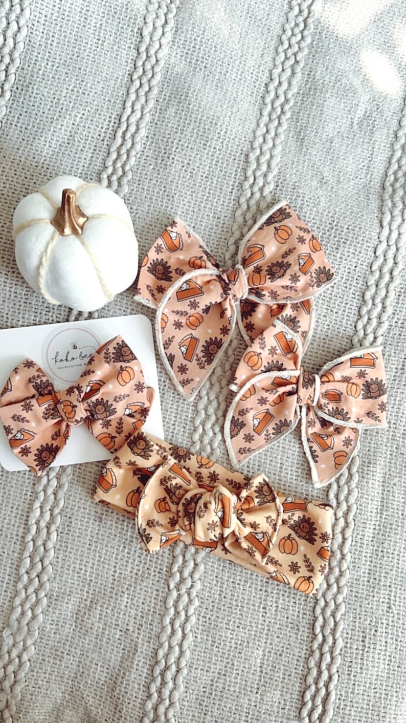 May include: Three brown and orange fabric hair bows with a pumpkin pie and turkey print. The bows are on a light gray knitted surface. There is a white pumpkin in the background.